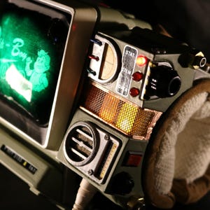 Op de afbeelding: Een gedetailleerde close-up van een Pip-Boy 3000 replica, een draagbaar apparaat met een klein scherm met groene tekst en een cartoonfiguur. Het apparaat is olijfgroen met diverse knoppen, draaiknoppen en een gewatteerde polsband.