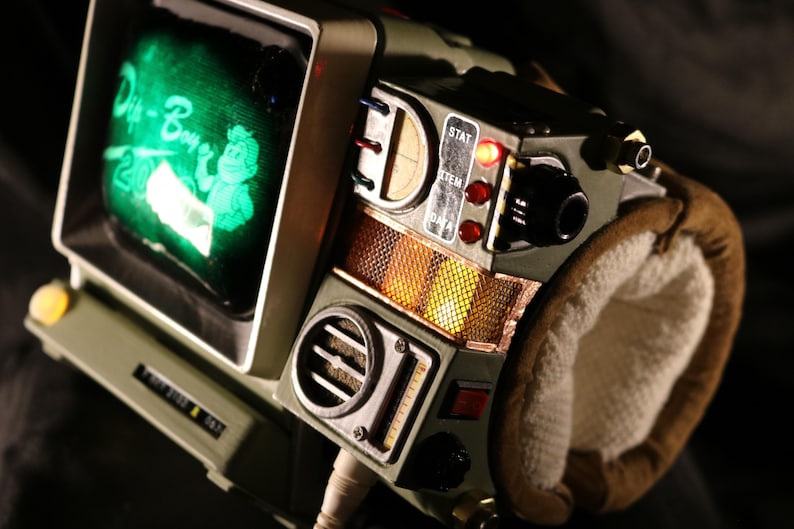 Op de afbeelding: Een gedetailleerde close-up van een Pip-Boy 3000 replica, een draagbaar apparaat met een klein scherm met groene tekst en een cartoonfiguur. Het apparaat is olijfgroen met diverse knoppen, draaiknoppen en een gewatteerde polsband.