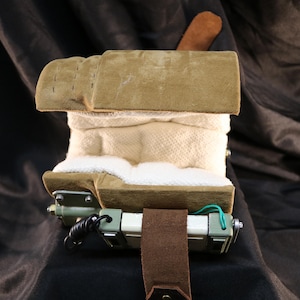 Op de afbeelding: Een complex apparaat met een buitenkant van bruin su&egrave;de, een witte stoffen binnenkant en een mechanische basis. Het apparaat heeft een bruine leren riem en een klein bruin leren stukje bovenop. De achtergrond is een donkere, getextureerde stof.