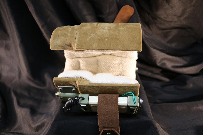 Op de afbeelding: Een complex apparaat met een buitenkant van bruin su&egrave;de, een witte stoffen binnenkant en een mechanische basis. Het apparaat heeft een bruine leren riem en een klein bruin leren stukje bovenop. De achtergrond is een donkere, getextureerde stof.