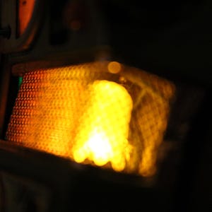Op de afbeelding: Close-up van een heldere, geel-oranje lichtbron, mogelijk een lamp of warmtebron, met een getextureerde, rasterachtige bekleding. Het licht is diffuus, tegen een donkere achtergrond.