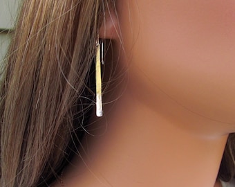 May include: A pair of long, thin, gold earrings with a hammered texture. The earrings are hanging from the earlobes of a person with long brown hair.