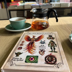 以下が含まれることがあります： 神話の生き物のイラストが描かれた「The Myth Hunter's Journal」というタイトルの本、緑色のティーカップとソーサー、木製のテーブルに紅茶が入ったガラスのティーポット。 本の表紙にはフェニックスが描かれています。