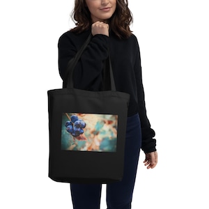 Puede incluir: Bolso tote negro con una imagen rectangular de uvas azul oscuro en una vid. El fondo de la imagen es una suave mezcla de verde, naranja y marrón. El bolso tote lo lleva una persona que lleva un suéter negro.