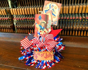 May include: A red, white, and blue Fourth of July themed centerpiece with a red, white, and blue bow, a red, white, and blue flag, and a red, white, and blue paper streamer. The centerpiece is on a wooden surface.