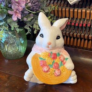 May include: A white ceramic bunny figurine with a pink scarf and a yellow basket filled with red and yellow flowers. The bunny has black eyes and a pink nose. The figurine is a decorative item.