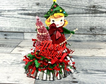 May include: A vintage Christmas tree topper featuring a paper doll dressed in red with a red bow and holding a green pipe cleaner. The doll is standing on a silver cupcake liner surrounded by red, green, and white shredded paper and a red "Merry Christmas" sign.