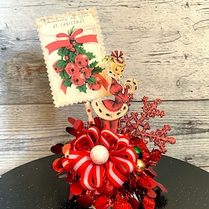 May include: A red and white Christmas centerpiece with a vintage postcard featuring a woman in a red dress and the text "Thinking of you at Christmas". The centerpiece also includes a red snowflake, a red and white bow, and red and green tinsel.