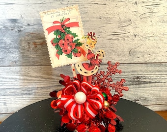 Puede incluir: Un centro de mesa navideño rojo y blanco con una postal vintage que presenta a una mujer con un vestido rojo y el texto "Thinking of you at Christmas". El centro de mesa también incluye un copo de nieve rojo, un lazo rojo y blanco y purpurina roja y verde.