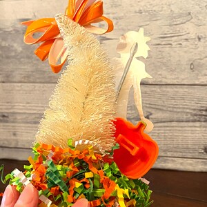 May include: A white bottle brush Christmas tree with an orange bow on top. The tree is decorated with green, orange, and yellow shredded paper. A silver silhouette of a person holding a guitar is behind the tree.