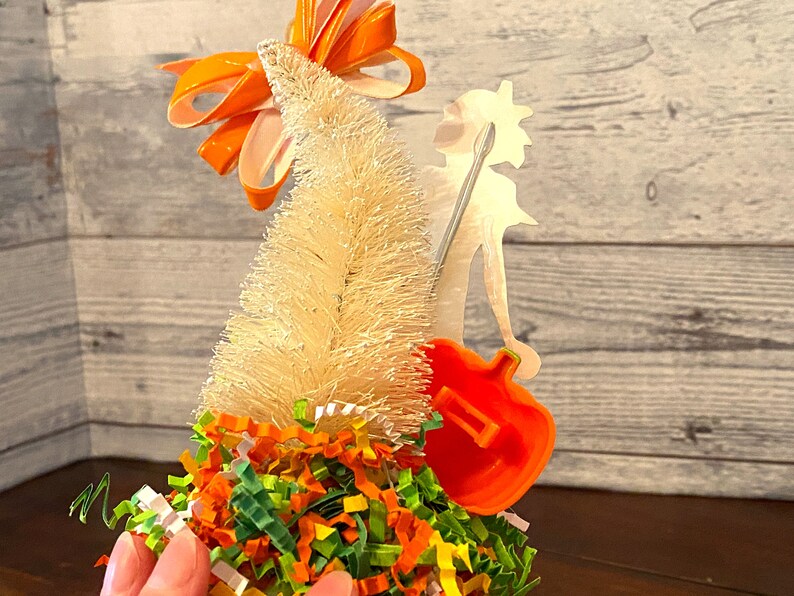 May include: A white bottle brush Christmas tree with an orange bow on top. The tree is decorated with green, orange, and yellow shredded paper. A silver silhouette of a person holding a guitar is behind the tree.