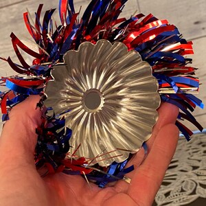 May include: A silver, fluted metal bundt pan is held in a hand, surrounded by red, white, and blue tinsel. The pan has a central hole and a decorative, scalloped edge. The tinsel adds a festive touch.