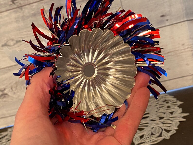 May include: A silver, fluted metal bundt pan is held in a hand, surrounded by red, white, and blue tinsel. The pan has a central hole and a decorative, scalloped edge. The tinsel adds a festive touch.