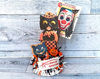 May include: A Halloween-themed centrepiece featuring a black cat with a witch's hat, a clown mask, and a pumpkin. The centrepiece is decorated with black and orange streamers and a sign that reads "Hallowe'en Greetings".