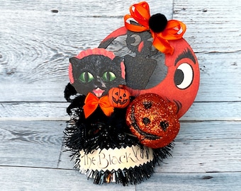 Könnte beinhalten: Eine Halloween-Dekoration mit einer schwarzen Katze, einem roten Halbmond mit einem Auge und einem glitzernden orangefarbenen Kürbis. Die Dekoration ist mit einer schwarzen und orangefarbenen Schleife und dem Text "The Black Cat" verziert.