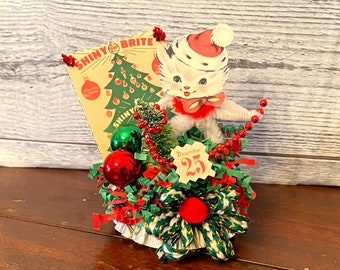 Puede incluir: Una decoración de mesa navideña con una figurita de gato blanco que lleva un gorro de Papá Noel, rodeada de adornos rojos y verdes, un pequeño árbol de Navidad y un letrero que dice "Shiny Brite".
