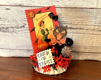Könnte beinhalten: Eine Halloween-Basteldekoration mit einer schwarzen Katze mit einer roten Tupfen-Schleife, einem Spinnennetz und einer Vintage-Postkarte mit einer Frau und einem Kürbis. Die Dekoration besteht aus roten und schwarzen Papierschnitzeln und einer silbernen Cupcake-Form. Die Postkarte lautet "Halloween Greetings". Die Dekoration enthält auch ein Schild mit der Aufschrift "Trick or Treat".