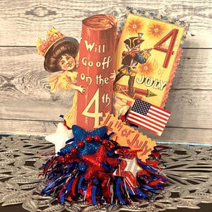 May include: A patriotic Fourth of July centerpiece featuring vintage-style illustrations. It includes a red firecracker, a card with a soldier, an American flag, and star-shaped decorations in red, white, and blue.