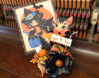 May include: A Halloween-themed decorative centerpiece featuring a square card with a witch illustration, a cat cutout holding a sign that reads Trick Or Treat, a small bottle brush tree, orange and black beads, and a large black ribbon bow.