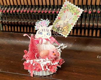 May include: A pink and white Easter figurine of a cat wearing a pink dress and a floral crown. The cat is holding a small pink Easter egg and a pink chick. The figurine is sitting in a nest of pink and white shredded paper.
