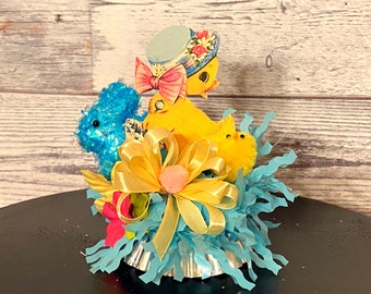 May include: A yellow chick wearing a blue hat with pink flowers and a pink bow sits in a blue nest with a yellow bow and a yellow chick with a fuzzy yellow body.