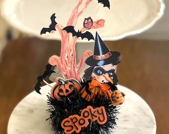May include: A Halloween-themed decoration featuring a witch with a black hat and mask, a spooky tree, and pumpkins. The decoration is surrounded by black tinsel and the word "Spooky" is written in black letters.