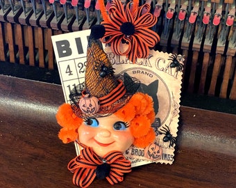 May include: A Halloween-themed craft featuring a doll's face with orange hair, a black witch's hat with a spider, and a black and orange bow. The craft is attached to a vintage "Black Cat" brand card with a black cat illustration and the text "Black Cat Brand".