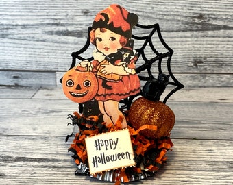 Könnte beinhalten: Eine Vintage-Halloween-Figur mit einem Mädchen in einem roten Kleid und Hut, das einen Kürbis mit Fratze hält. Die Figur ist umgeben von schwarzen Spinnweben, einer schwarzen Katze und orange-schwarzem zerfetzten Papier. Das Schild lautet "Happy Halloween".