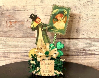 May include: A St. Patrick's Day greeting centerpiece with a green, gold, and white color scheme. It features a paper cutout of a man wearing a green suit and top hat, a paper cutout of a woman wearing a green hat, a gold coin, green shamrocks, and a sign that reads "St. Patrick's Day Greeting".