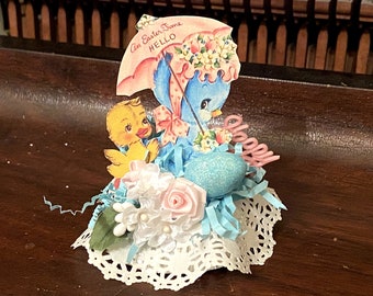 Pode incluir: Enfeite de Páscoa com um pássaro azul sob um guarda-chuva e um pintinho amarelo. Inclui um ovo azul, rosas cor-de-rosa e uma base de renda. O texto diz "An Easter Time HELLO".