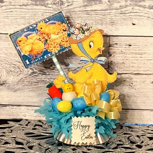 May include: A yellow chick figurine with a blue bow and a hat with flowers, holding a blue and white striped stick with a vintage postage stamp attached. The chick is surrounded by blue and yellow Easter eggs, a yellow bow, and blue tissue paper. The base of the figurine is a silver cupcake liner with the words "Happy Easter" printed on it.