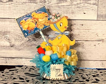 May include: A yellow chick figurine with a blue bow and a hat with flowers, holding a blue and white striped stick with a vintage postage stamp attached. The chick is surrounded by blue and yellow Easter eggs, a yellow bow, and blue tissue paper. The base of the figurine is a silver cupcake liner with the words "Happy Easter" printed on it.