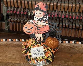 Könnte beinhalten: Ein Halloween-Cupcake-Topper mit einer Vintage-Illustration eines Kindes im orangefarbenen Kostüm, das einen Kürbis und eine schwarze Katze hält. Er enthält einen glitzernden orangefarbenen Kürbis, ein schwarzes Spinnennetz und ein Happy Halloween-Schild.