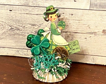 May include: A green, white, and gold St. Patrick's Day themed centerpiece with a paper doll wearing a green dress and hat, shamrock embellishments, and a ticket that reads "Patrick's Day Elf & Cabbie-ee", "Town Hall", "Admit 1".