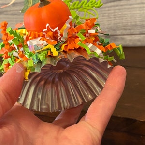 May include: A small, silver-colored metal pumpkin-shaped mold with a ruffled edge. The mold is decorated with colorful paper shreds and a small orange pumpkin.