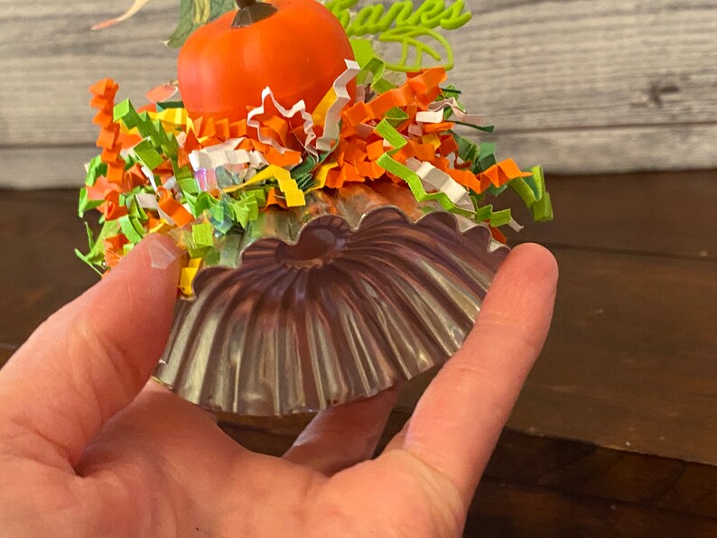 May include: A small, silver-colored metal pumpkin-shaped mold with a ruffled edge. The mold is decorated with colorful paper shreds and a small orange pumpkin.