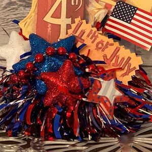 May include: A patriotic Fourth of July decoration featuring red, white, and blue glittery stars, red bead garland, and an American flag. The decoration includes a tag that reads "4th of July".