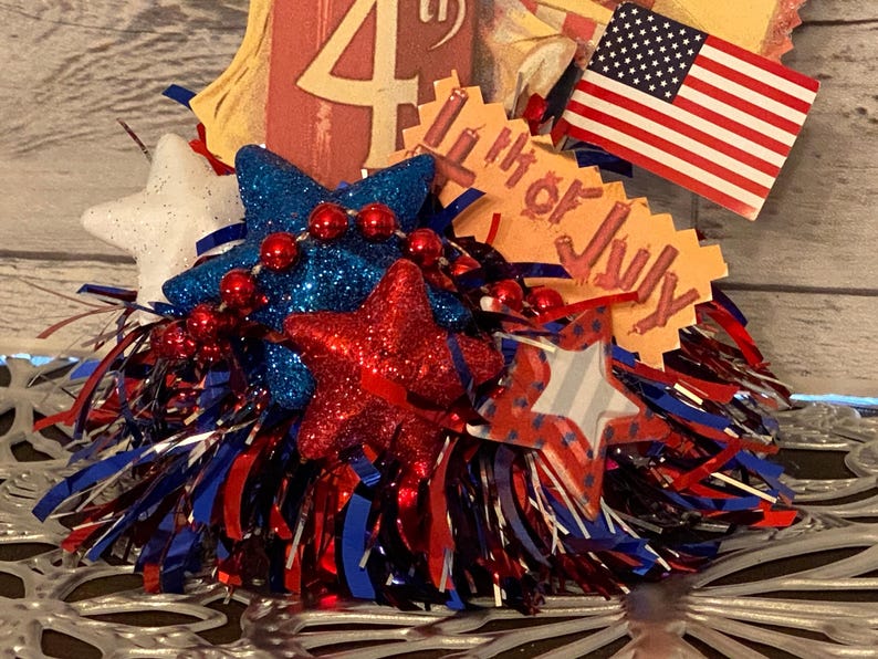 May include: A patriotic Fourth of July decoration featuring red, white, and blue glittery stars, red bead garland, and an American flag. The decoration includes a tag that reads "4th of July".