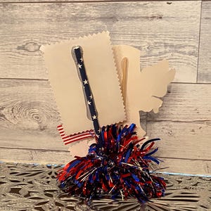May include: A patriotic-themed craft item featuring a blue straw with white stars, a red, white, and blue pom-pom, and a decorative paper cutout. The design incorporates red, white, and blue ribbon accents, evoking a festive atmosphere.