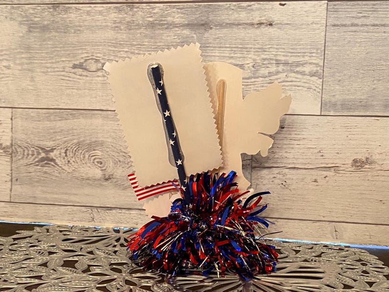 May include: A patriotic-themed craft item featuring a blue straw with white stars, a red, white, and blue pom-pom, and a decorative paper cutout. The design incorporates red, white, and blue ribbon accents, evoking a festive atmosphere.