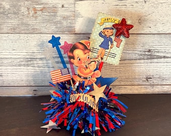 May include: A patriotic Fourth of July centerpiece with red, white, and blue tinsel, star-shaped accents, and vintage-style illustrations. Features a child playing a trumpet and a girl saluting with a flag. The words "OLD GLORY" are visible.