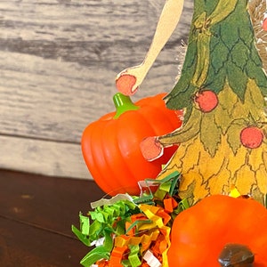 May include: A fall-themed craft featuring a paper cutout of a woman in a green and yellow dress holding a small orange pumpkin. The woman is standing on a bed of colorful shredded paper with two additional orange pumpkins.