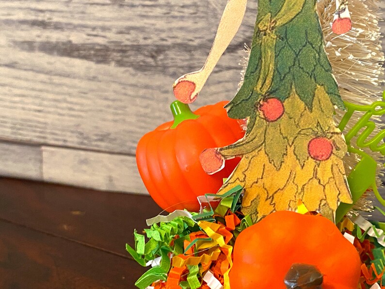 May include: A fall-themed craft featuring a paper cutout of a woman in a green and yellow dress holding a small orange pumpkin. The woman is standing on a bed of colorful shredded paper with two additional orange pumpkins.