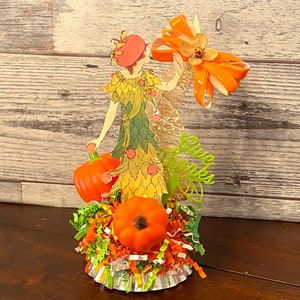 May include: A vintage-style paper doll figurine dressed in a green and yellow dress with a red hat, holding a small orange pumpkin. The doll is surrounded by orange and green shredded paper and a large orange bow. The text "Give Thanks" is written in green letters on the side of the figurine.