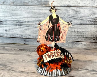 May include: A vintage-inspired Halloween decoration featuring a paper doll dressed in a black dress with a crescent moon and stars on her cape. The doll is standing on a silver base with a black spider, orange and black ribbon, and a sign that says "Trick or Treat".