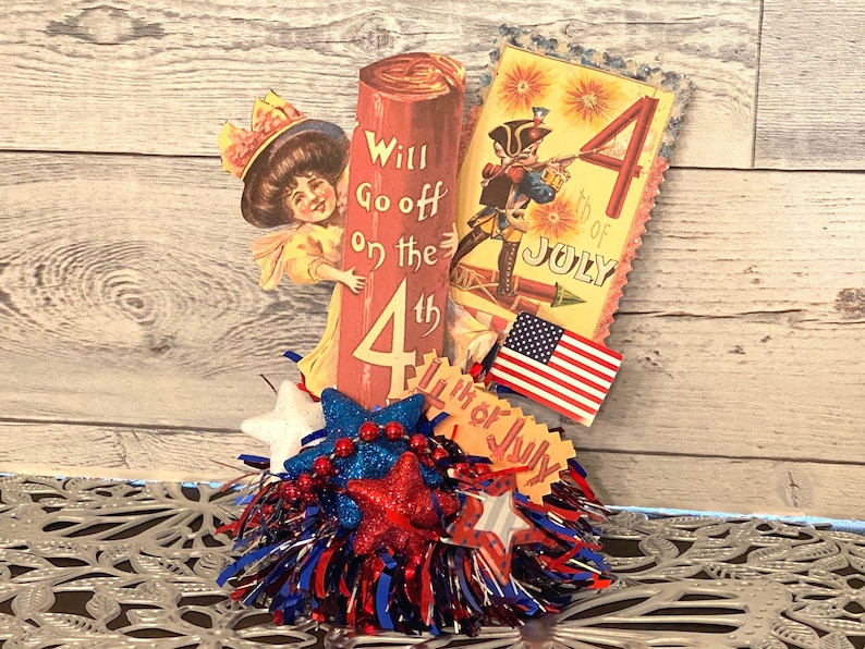 May include: A patriotic Fourth of July centerpiece featuring vintage-style illustrations. Includes a woman peeking from behind a firecracker, a soldier, an American flag, and star-shaped decorations in red, white, and blue.