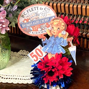 May include: A vintage-inspired centerpiece featuring a red, white, and blue color scheme. A paper doll with a blue and white dress holds a white umbrella. The doll is standing on a base of red and white flowers with blue and white tinsel. A sign reads "50¢". A vintage label for "P.C. Flett & Co's Rhubarb & Gooseberry Jam" is also included.