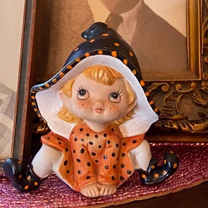 May include: A ceramic figurine of a blonde elf wearing a black witch hat with orange polka dots and a matching orange dress with black polka dots. The elf has large eyes, freckles, and black pointed shoes with orange polka dots, sitting in a pose.