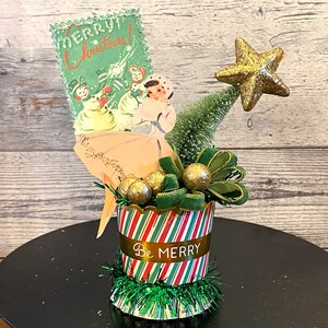 May include: A vintage Christmas decoration featuring a green and red striped container with the words "Be Merry". It includes a Christmas card with children, a bottle brush tree, gold star, and gold ornaments.