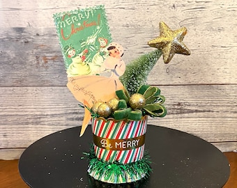 Puede incluir: Decoración navideña vintage con un recipiente a rayas verdes y rojas con la inscripción «Be Merry». Incluye una tarjeta navideña con niños, un árbol de cepillo de botella, una estrella dorada y adornos dorados.